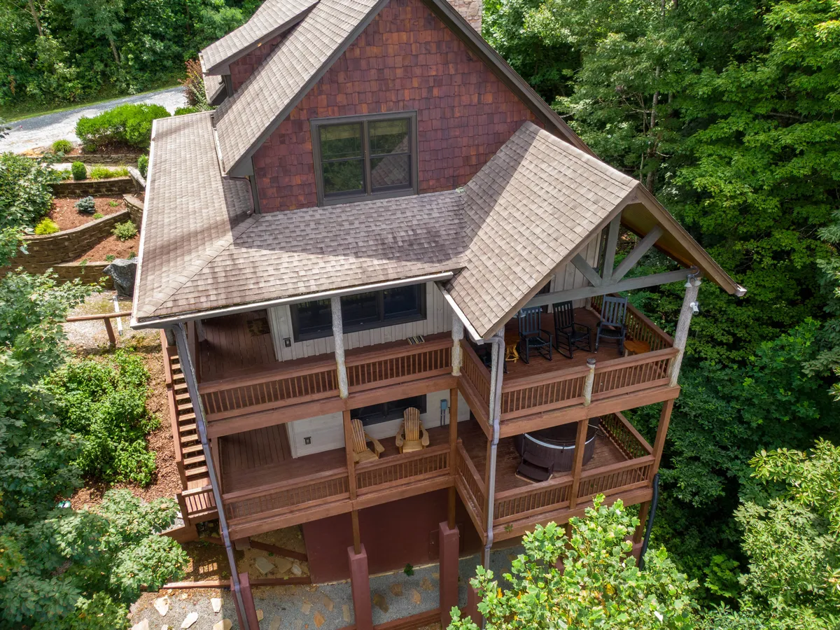 A wide, multi-level wooden deck surrounded by lush greenery, featuring rocking chairs for relaxation and a stone pathway leading into a forested backdrop. The deck is spacious, inviting families to gather for meals or conversations, with plenty of room for children to play or adults to unwind. This exterior space visually connects the cabin to its mountain environment, emphasizing both togetherness and exploration.