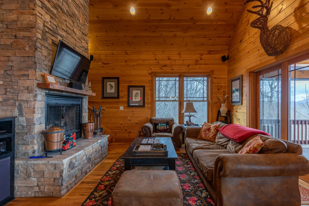 A warmly lit living room featuring a striking stone fireplace, plush leather sofas, rustic wooden walls, and a decorative antler. Sunlight filters through large windows, emphasizing the cozy seating and inviting atmosphere. The windows frame glimpses of the surrounding mountain scenery, merging the indoors with nature.
