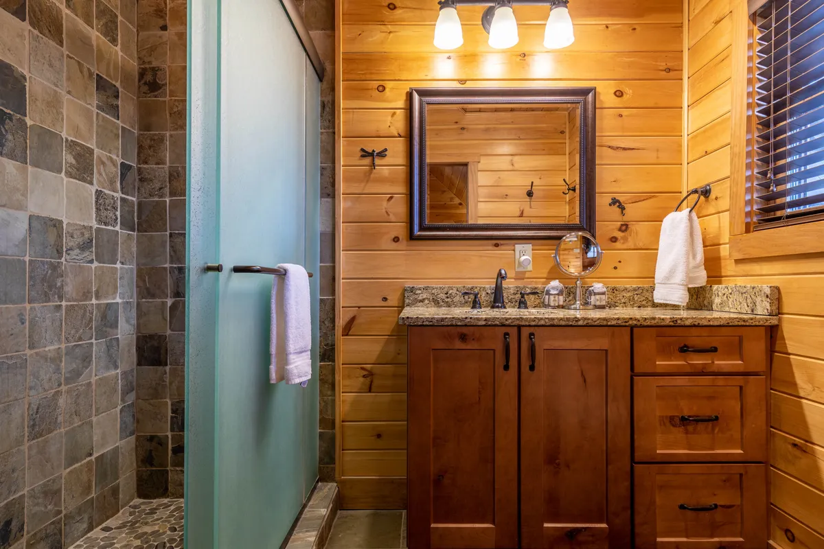 A serene bathroom featuring a granite countertop, wooden cabinetry, slate shower wall, framed mirror, and modern fixtures. The soothing color palette and natural materials evoke a spa-like environment ideal for refreshing showers or calming evening rituals.