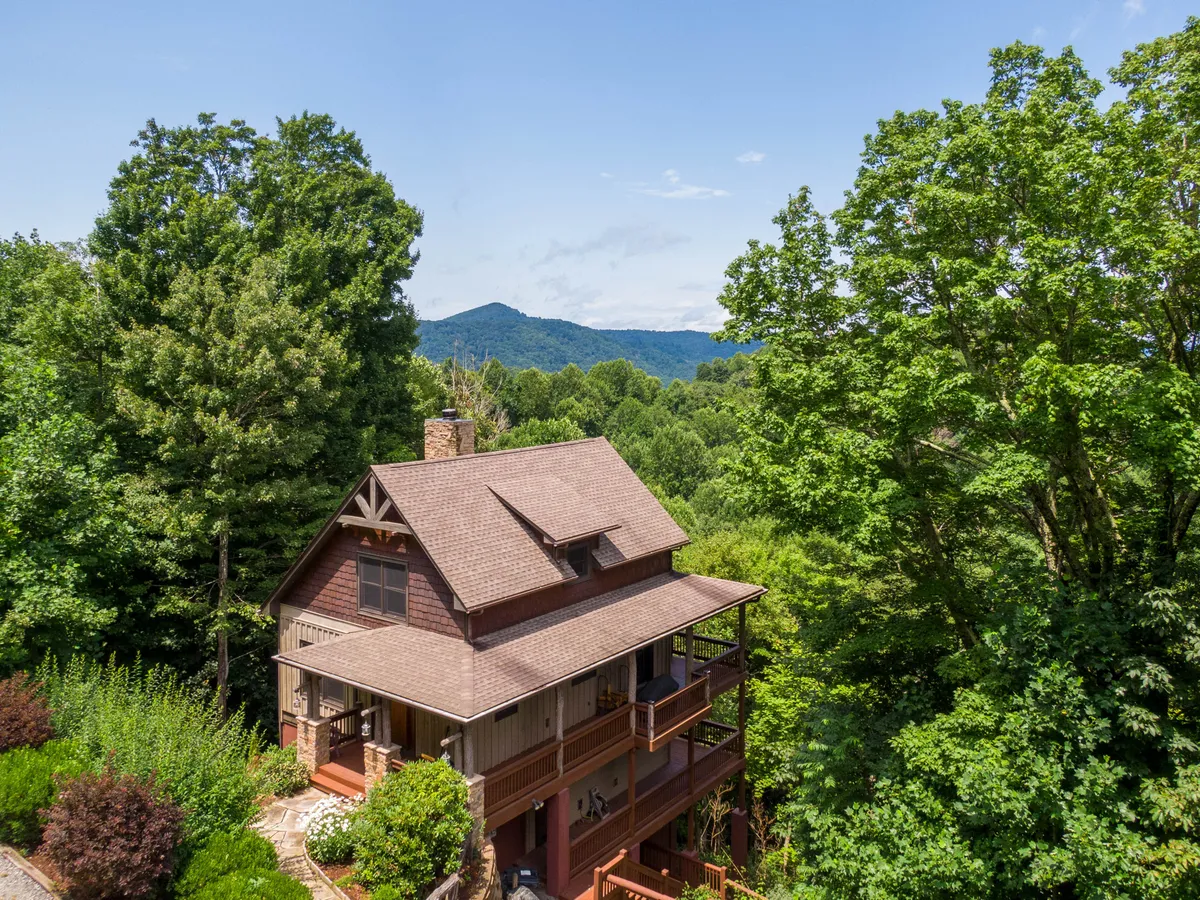 The hero image features a beautifully crafted Adirondack-style cabin nestled among towering trees, set against a breathtaking mountain backdrop. The spacious porch is perfect for relaxing while soaking in panoramic sunset views, and the inviting setting hints at the opportunity for mornings with coffee in the crisp mountain air or evenings gathered around a fire pit with loved ones.