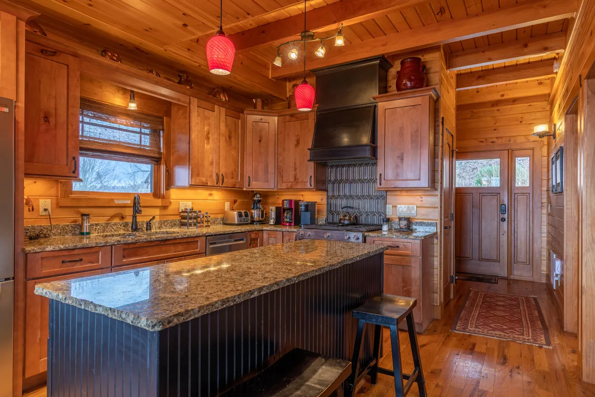 A warmly lit kitchen featuring chiseled edge granite countertops, custom wooden cabinetry, a large kitchen island topped with red pendant lights, and modern stainless steel appliances. The space exudes both rustic charm and upscale amenities, ideal for cooking and social gatherings.