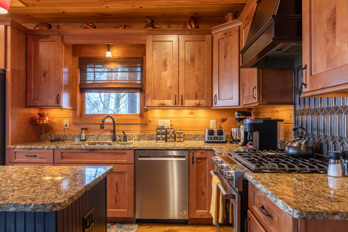 A cozy kitchen space with granite countertops, warm wood cabinetry, stainless steel appliances, and a window overlooking natural scenery. A kettle sits on the stove, highlighting the area’s functionality and inviting ambiance for meal preparation and casual family gatherings.