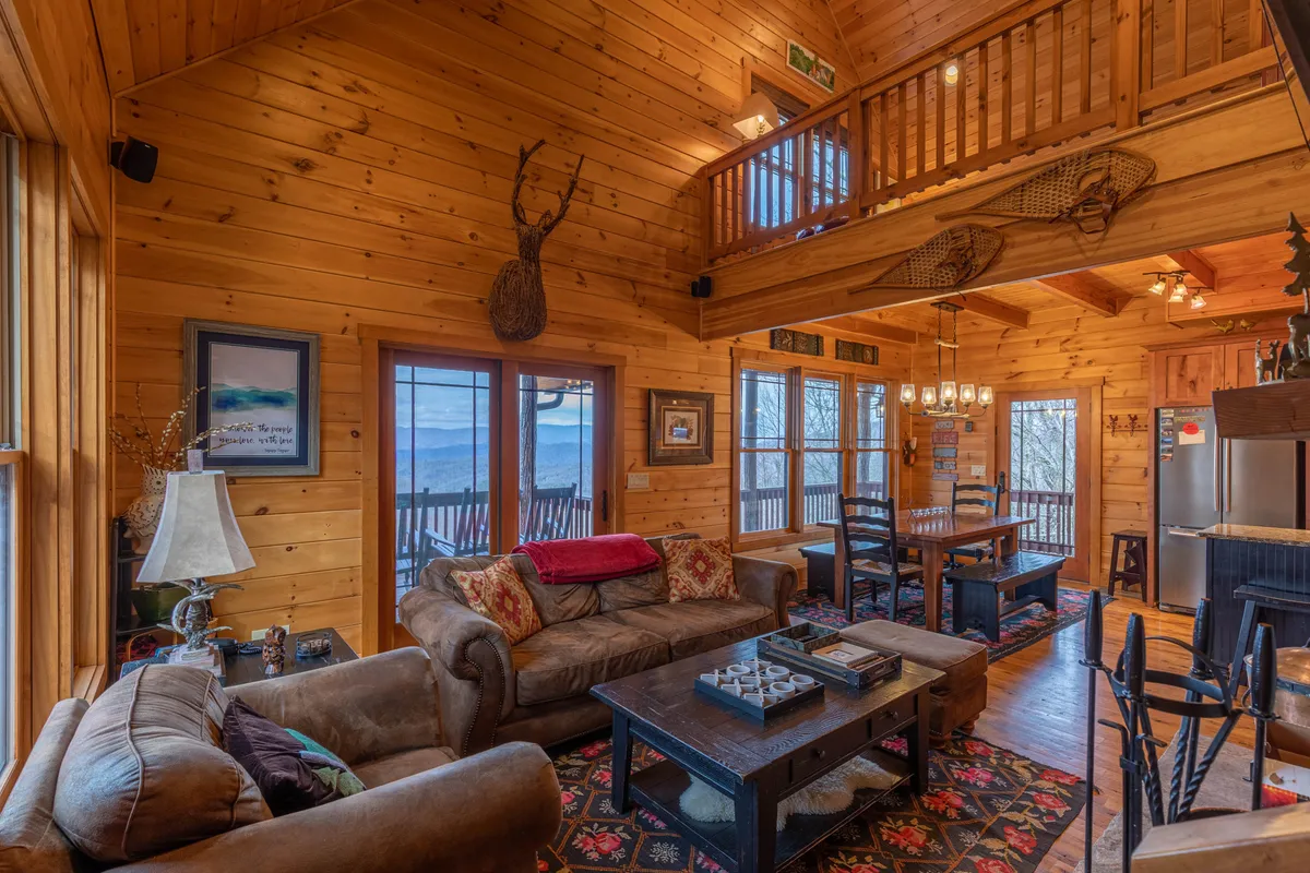 An open-concept living and dining space with exposed wooden beams, a comfortable sofa, a welcoming dining table, and expansive windows showcasing nature outside. The room is bathed in warm, inviting light, and the space is arranged for entertaining and relaxation.