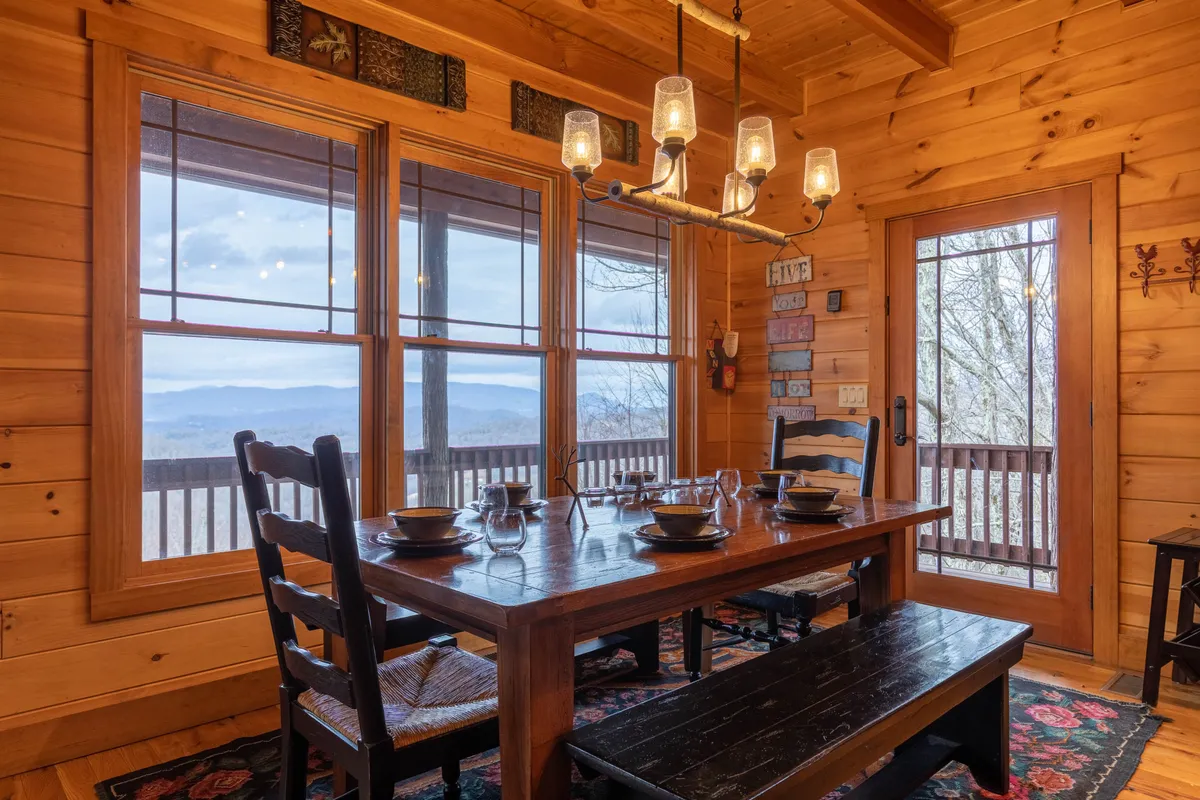 A spacious dining area with a long wooden table set beneath a sparkling chandelier. Large windows flood the room with natural light and offer views of the mountains beyond. Decorative wall art adds charm, while the setup suggests space for group celebrations and shared meals.