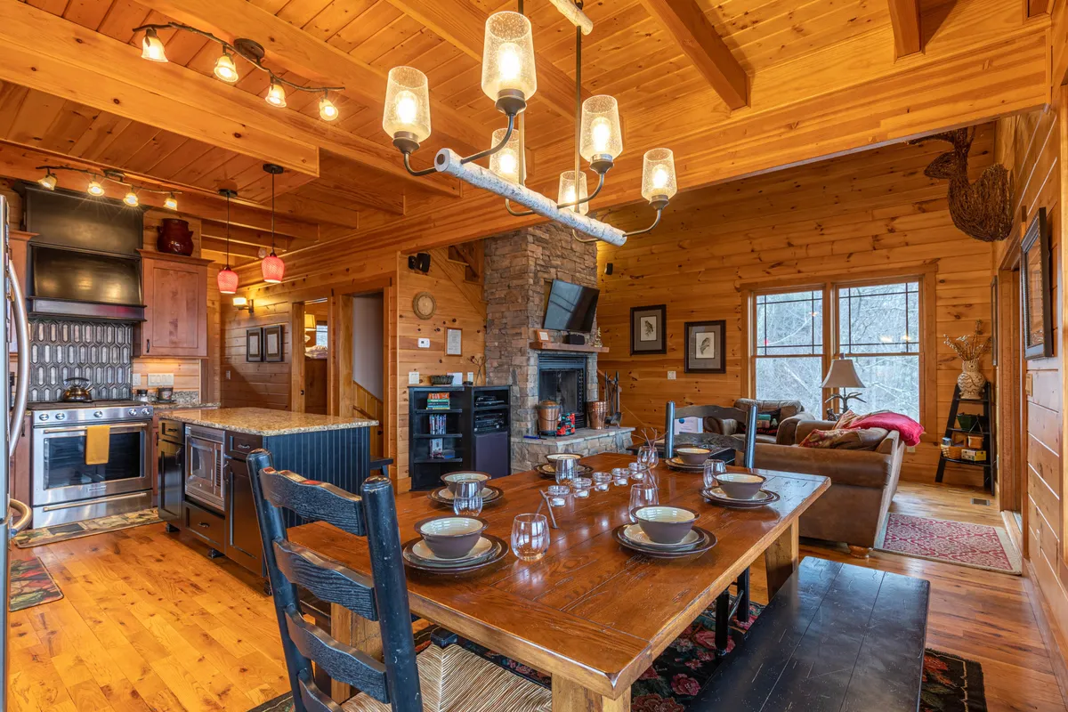 A spacious dining area set with a wooden table, elegant place settings, and views of both the stone fireplace and nearby kitchen appliances. Modern light fixtures and the warm glow of the fireplace create an atmosphere perfect for sharing meals and laughter with loved ones.