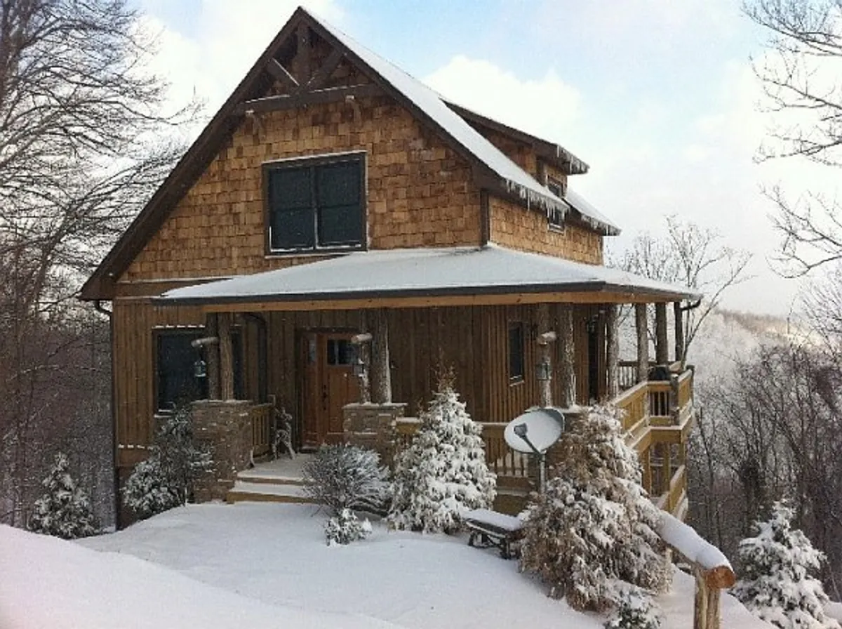 The cabin’s exterior shown with a snow-covered roof and surrounding trees. The front porch is visible, suggesting cozy indoor-outdoor transitions and seasonal enjoyment. The warm wooden structure stands out against the winter backdrop, underscoring both the safety and comfort of the retreat in all seasons.