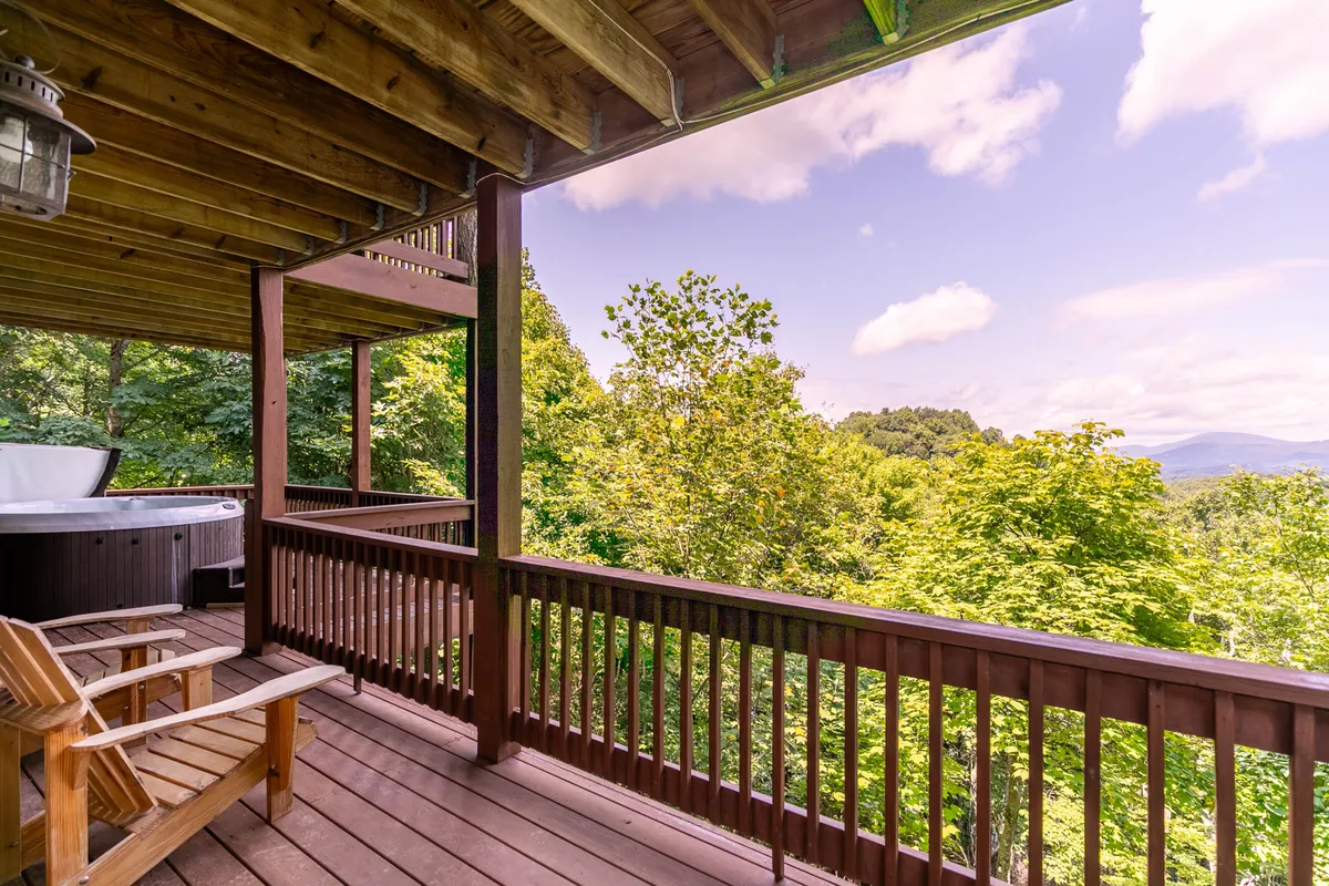 A serene exterior view of a wooden deck featuring a private hot tub, surrounded by lush trees and offering a sweeping mountain vista. The deck is positioned to maximize views of both sunrise and the night sky, making it ideal for stargazing or soaking in the hot tub.