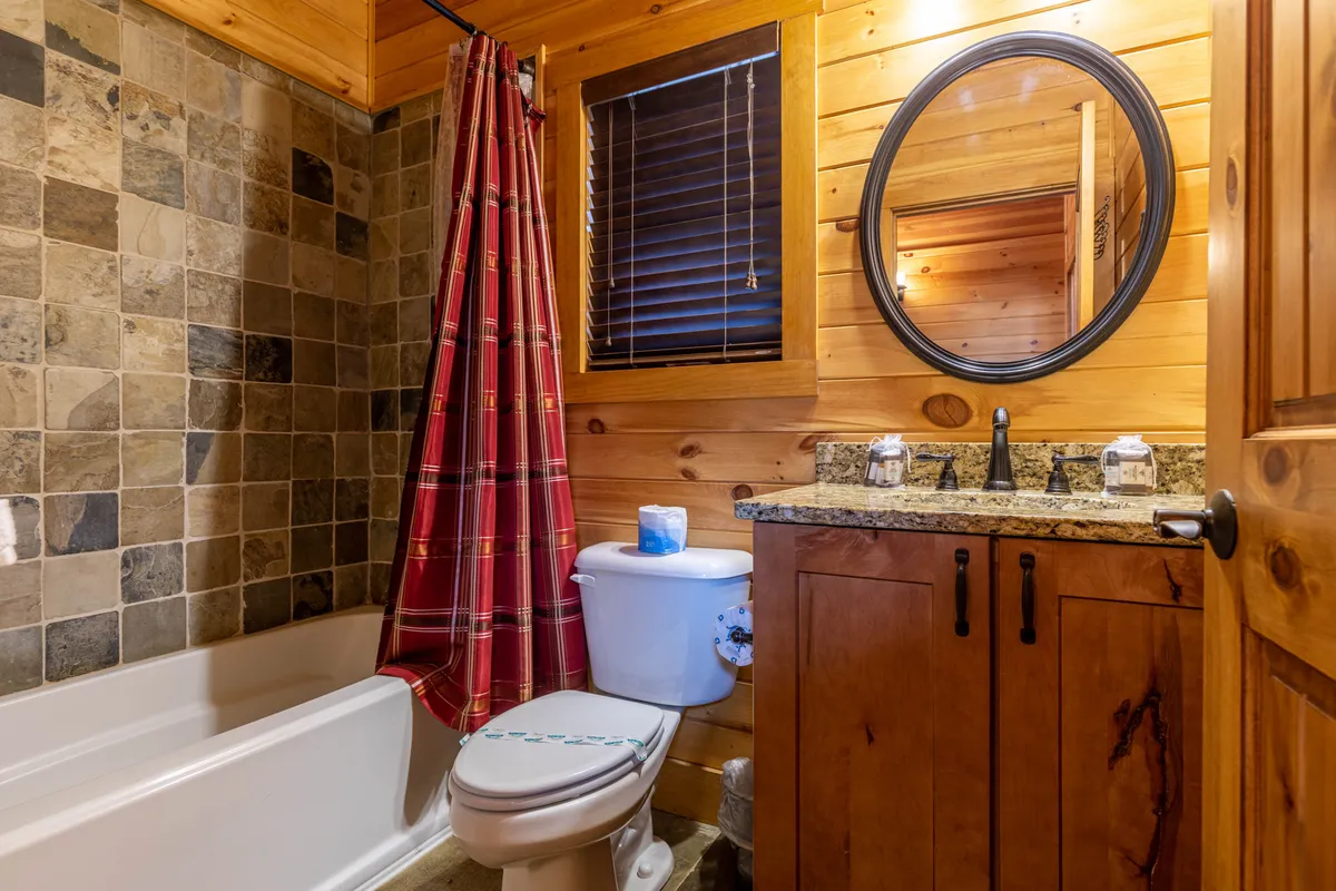 A rustic bathroom with a granite countertop, plaid shower curtain, wood-paneled walls, and a round mirror. The inviting space hints at relaxing baths and moments of self-care after a day outdoors, with warm wood tones and thoughtful, homey touches.