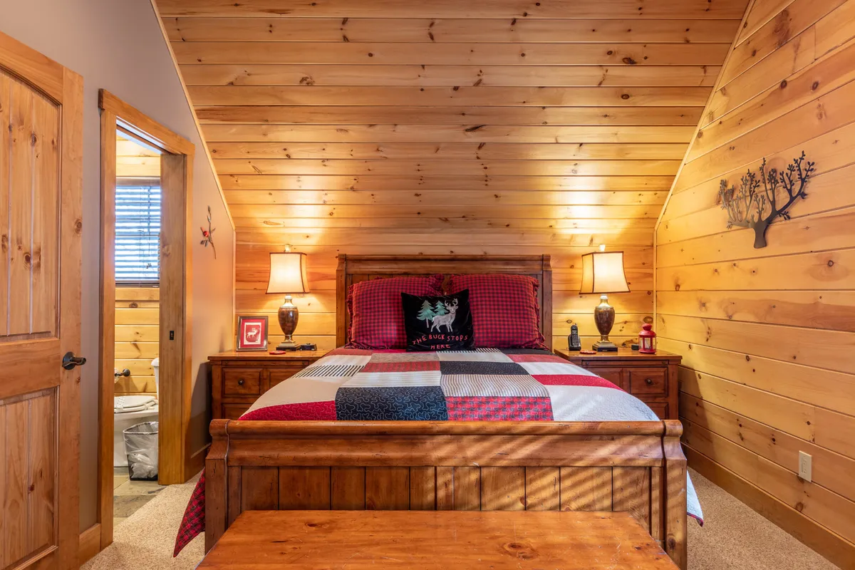 A welcoming bedroom with a wooden bed dressed in plaid bedding, warm bedside lamps, and classic wooden furniture. The room’s soft lighting and inviting textures create a restful, cozy environment ideal for relaxing or reading.